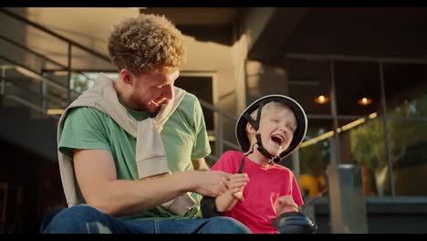 Happy-young-dad-in-a-green-t-shirt-with-curly-hair-tickles-his-little-son-in-a-red-t-shirt-and-a-black-helmet-near-a-large-building-on-the-street.-Happy-son-and-father-spending-time-in-the-park-while-riding-a-skateboard