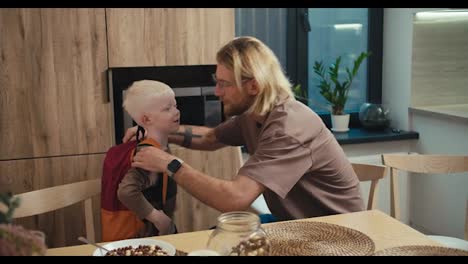 A-blond-man-in-glasses-with-a-beard-hugs-his-little-albino-boy-with-white-hair-who-put-on-a-red-backpack-before-his-day-at-school.-Happy-little-boy-says-goodbye-to-his-father-and-goes-to-school