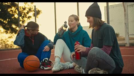 A-trio-of-blonde-girls-in-sportswear-drink-water-from-sports-bottles-and-chat-during-a-break-from-basketball-practice-near-an-orange-ball-on-a-red-street-court-at-sunrise