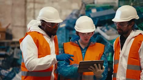 A-brunette-girl-in-a-blue-uniform-and-a-White-protective-helmet-in-an-orange-vest-holds-a-tablet-in-her-hands-and-tells-her-fellow-guys-about-her-plans-at-the-plastic-and-waste-recycling-plant