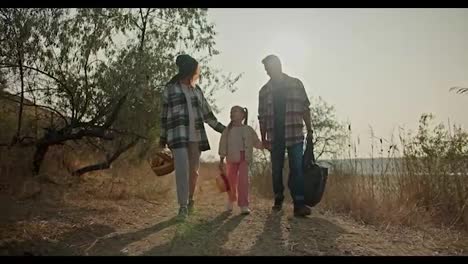 A-brunette-girl-in-a-Green-hat-in-a-Green-checkered-shirt,-along-with-a-basket,-walks-hand-in-hand-with-her-little-daughter-and-her-husband,-a-man-with-gray-hair-in-a-checkered-shirt,-on-a-picnic-in-the-summer