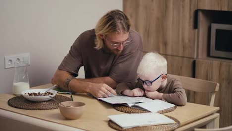 Ein-Glücklicher-Albino-Junge-Mit-Weißem-Haar-Und-Runder-Blauer-Brille-Lernt-In-Der-Schule-Mit-Hilfe-Spezieller-Lehrbücher-Lesen-Und-Schreiben-Und-Sein-Blonder-Vater-Hilft-Ihm-In-Einer-Modernen-Küche-Bei-Den-Hausaufgaben