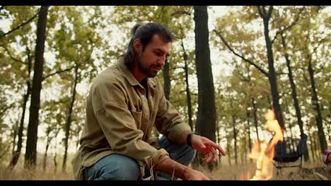 A-brunette-man-in-a-light-green-jacket-puts-firewood-in-a-small-fire-during-a-halt-on-a-hike-in-a-yellow-green-autumn-forest-with-withered-grass.-Happy-man-resting-during-a-rest-near-a-fire-while-hiking-in-the-forest