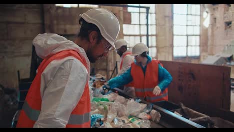 A-brunette-man-in-a-special-uniform-and-an-orange-vest-recycles-garbage-and-selects-plastic-bottles-of-a-specific-color-along-with-his-employees-near-a-conveyor-belt-at-a-waste-recycling-plant