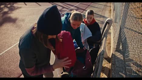 A-trio-of-girls-in-sportswear-put-their-things-in-a-backpack-on-a-street-sports-field-after-playing-basketball-on-a-sunny-summer-morning.-Three-blonde-girls-in-sports-summer-uniforms-collect-their-things-after-playing-basketball