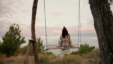 Close-up-shooting-from-behind:-A-brunette-girl-in-a-checkered-shirt-swings-on-a-wooden-swing-outside-the-city.-Rest-in-the-country-by-the-sea