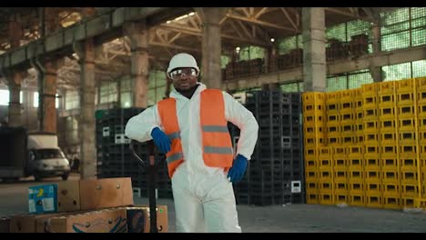 Portrait-of-a-confident-man-with-Black-skin-in-a-white-protective-uniform-and-an-orange-vest-who-stands-in-safety-glasses-near-large-boxes-for-sorted-waste-at-a-plant-for-processing-and-sorting-garbage-and-waste