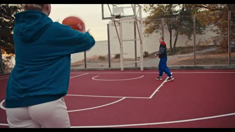 A-blonde-girl-in-white-pants-throws-a-basketball-to-her-friend-who-throws-it-into-the-basket-on-a-red-colored-basketball-field-early-in-the-morning-at-sunrise