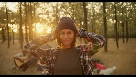 Portrait-of-a-happy-girl-with-a-bob-hairstyle-in-a-black-hat-in-a-plaid-shirt-against-the-backdrop-of-hikers-and-tents-in-a-Sunny-summer-green-forest.-Happy-woman-posing-and-looking-at-camera-while-hiking