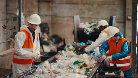 Shot-from-the-side-three-workers-in-white-uniforms-in-orange-vests-sort-garbage-near-a-conveyor-belt-transferring-plastic-bottles-by-color-into-the-appropriate-boxes-at-a-waste-recycling-plant