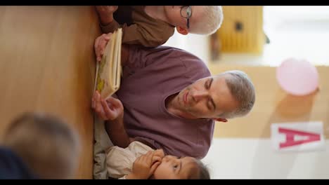 Vertical-video-of-a-man-with-gray-hair-in-a-purple-T-shirt-reading-a-book-to-children-who-are-lying-on-the-floor-on-special-pillows-and-listening-to-him-in-a-club-for-preparing-children-for-school