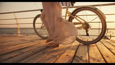 Close-up-shot-of-a-girl-in-a-long-white-light-dress-who-wriggles-in-the-wind-and-walks-near-her-bicycle-along-the-beach,-which-is-covered-with-boards-along-the-sea-at-Sunrise-in-summer