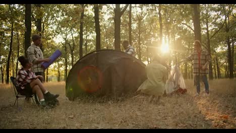 A-happy-company-of-6-hiking-participants-in-hiking-clothes-lay-out-their-things-and-set-up-a-large-black-tent-for-an-overnight-stay-in-the-forest-during-the-summer-during-the-evening