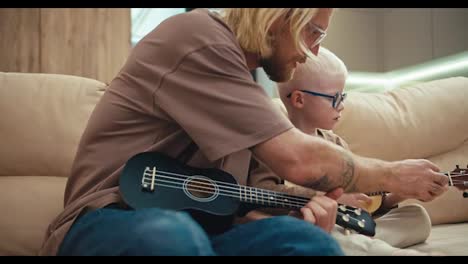 Happy-blond-man-with-a-beard-and-glasses-teaches-his-little-albino-son-in-blue-glasses-to-play-the-guitar-correctly-and-press-chords-using-the-fingers-of-his-left-hand-on-the-sofa-at-home