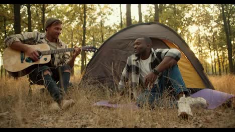 A-happy-man-with-black-skin-in-a-checkered-shirt-sings-and-a-blond-man-in-a-hat-plays-the-guitar-against-of-a-gray-tent-in-a-green-forest-during-a-summer-halt.-A-happy-company-of-two-guys-during-a-rest-in-a-sunny-light-green-summer-forest