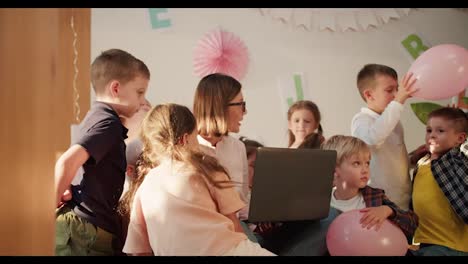 A-girl-teacher-with-a-bob-hairstyle-and-glasses-holds-a-gray-laptop-in-front-of-her-and-communicates-with-children-in-the-room-for-preparing-for-school-in-a-club-for-preschool-children.-Lesson-at-the-club-to-prepare-children-for-school