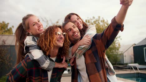 Two-girls-are-sitting-on-their-boyfriends'-backs-in-checkered-shirts.-A-guy-with-long-hair-takes-a-funny-selfie-near-the-sunbeds.-Rest-in-the-country-house