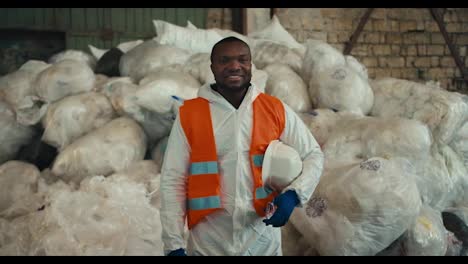 Portrait-of-a-happy-and-confident-brunette-man-with-Black-skin-in-a-white-protective-uniform-and-an-orange-vest-who-stands-near-a-pile-of-recycled-and-sorted-cellophane-at-a-waste-processing-and-sorting-plant