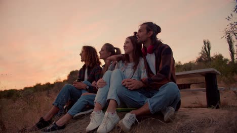 Two-couples-in-love-are-sitting-in-nature.-Two-guys-are-pointing-their-hands-forward-and-talking.-Rest-outside-the-city-against-the-background-of-the-sunset.-Rest-in-the-country-house