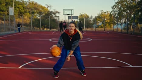 Porträt-Eines-Blonden-Mädchens-In-Sportkleidung-Und-Blauen-Hosen,-Das-Einen-Orangefarbenen-Basketballball-Vom-Boden-Abprallen-Lässt,-Damit-Manövriert-Und-Ihn-Dann-Im-Sommer-Auf-Einem-Roten-Basketballplatz-Auf-Der-Straße-In-Den-Korb-Wirft