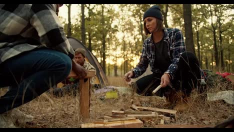 A-girl-in-a-black-hat-with-a-bob-hairstyle-helps-a-man-with-Black-skin-in-a-checkered-shirt-chop-wood-with-a-hunting-knife-during-a-camping-stop-in-a-sunny-summer-green-forest