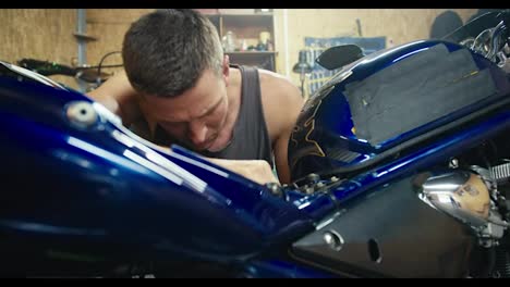 Close-up-shot:-a-man-in-a-gray-T-shirt,-a-mechanic-with-a-short-haircut,-is-repairing-his-gray-motorcycle-in-his-workshop---a-garage.-Inspection-of-equipment-in-the-workshop