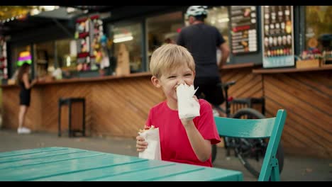 Happy-little-blond-boy-in-a-red-T-shirt-sits-at-a-table-at-a-street-cafe-and-eats-a-hot-dog.-Little-boy-eating-hot-dogs-in-a-cafe-in-the-park