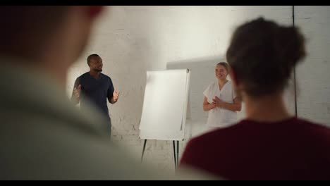 Over-the-shoulder:-A-Black-male-doctor-in-a-blue-uniform,-and-his-nurse-assistant-in-a-white-uniform-conduct-a-training-session-against-a-white-brick-wall-for-an-audience-that-is-listening