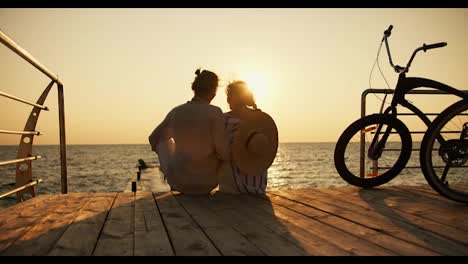 A-happy-couple,-a-guy-and-a-girl,-are-sitting-on-the-shore-of-a-beach-that-is-covered-with-boards-and-looking-at-the-sunrise-on-the-sea,-their-bike-is-located-near-them.-Happy-date-on-the-beach-near-the-sea-at-sunrise