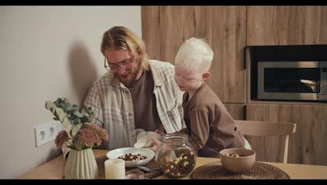 A-little-albino-boy-with-white-hair-and-blue-eyes-pours-milk-into-a-plate-for-his-father,-a-blond-man-with-a-beard-and-glasses-in-a-white-checkered-shirt-during-their-breakfast-in-a-modern-kitchen