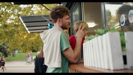 A-father-with-curly-hair-in-a-green-T-shirt-and-his-son,-a-little-blond-boy-in-a-red-T-shirt,-make-an-order-in-a-street-cafe-in-the-park