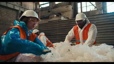 A-brunette-girl-in-a-blue-uniform-and-a-white-protective-helmet-together-with-her-male-colleagues-in-orange-vests-sorts-and-draws-cellophane-garbage-at-a-waste-processing-and-sorting-plant