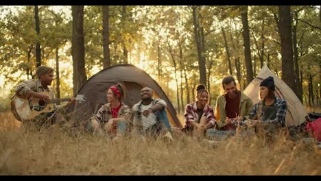 A-company-of-6-trekking-participants-in-special-hiking-clothes-against-the-backdrop-of-tents.-A-blond-man-in-a-hat-plays-an-acoustic-guitar-and-all-the-other-participants-in-the-hike-sing-along-and-palm-him-off-against-the-backdrop-of-a-green,-sunny-summer-forest