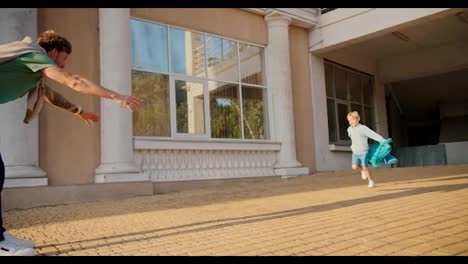 A-happy-little-blond-boy-in-blue-clothes-runs-up-to-his-father-along-with-his-backpack-and-jumps-into-his-arms.-Dad-meets-his-little-son-after-school