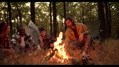 A-brunette-man-in-a-light-green-jacket-adjusts-firewood-in-a-fire-while-other-participants-in-a-group-hike-sit-and-enjoy-the-views-of-bright-flames,-fry-marshmallows-and-have-fun-in-the-forest