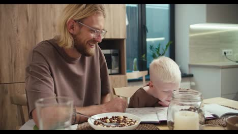 A-little-albino-boy-with-white-hair-reads-a-book-with-his-father,-a-blond-man-with-glasses-and-a-beard,-in-the-kitchen-after-breakfast-in-the-morning-in-a-modern-apartment