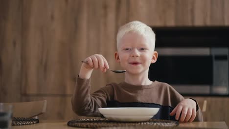 Portrait-of-a-happy-albino-boy-with-white-hair-who-has-breakfast-in-the-kitchen-and-eats-oatmeal-with-a-spoon-while-sitting-at-a-modern-table-in-a-modern-kitchen-at-home