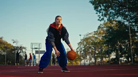 Portrait-of-a-blonde-girl-in-a-sports-uniform-and-blue-pants-who-bounces-an-orange-basketball-ball-off-the-floor-and-throws-it-into-the-hoop-on-a-red-street-court-in-the-summer