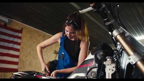 A-tired-brunette-girl-mechanic-finishes-her-work-and-repairs-her-bike.-Girl-working-in-a-workshop-with-tools-and-USA-flag.-End-of-a-hard-day-at-work-in-the-workshop