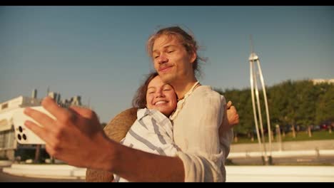 Un-Chico-Sostiene-Un-Teléfono-En-Sus-Manos-Y-Se-Toma-Una-Selfie-Con-Su-Novia,-Quien-Lo-Besa-En-La-Mejilla-En-Una-Playa-Moderna-En-El-Verano-Al-Amanecer.-Chico-Y-Chica-Felices-En-Una-Cita-Tomándose-Una-Selfie