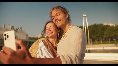 Ein-Glückliches-Paar,-Ein-Mann-Und-Eine-Frau-Machen-Ein-Selfie-Mit-Einem-Weißen-Telefon-An-Einem-Modernen-Strand.-Ein-Glückliches-Date,-Ein-Mann-In-Einem-Weißen-Hemd-Und-Ein-Braunhaariges-Mädchen-In-Einem-Weiß-blauen-Hemd-Machen-Ein-Selfie-Mit-Einem-Weißen-Telefon-An-Einem-Modernen-Strand.