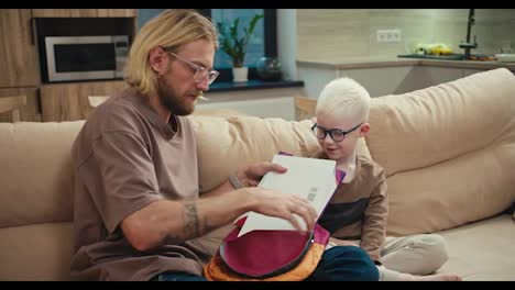 A-blond-man-in-glasses-with-a-beard-helps-his-little-son,-an-albino-boy-with-white-hair-color-in-blue-glasses,-collect-his-bright-briefcase-for-school-while-preparing-his-homework-at-home-in-a-modern-apartment