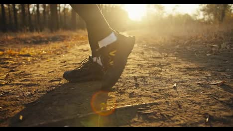 Close-up,-a-man-in-a-black-sports-uniform-and-black-sneakers-stretches-his-feet-before-starting-to-jog-along-an-earthen-path-in-the-autumn-forest-at-Sunrise-on-a-sunny-autumn-day