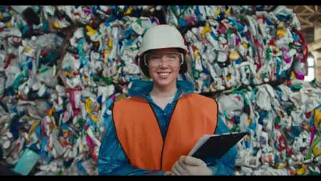 Close-up-portrait-of-a-brunette-girl-in-a-blue-protective-uniform-and-an-orange-vest-standing-against-the-backdrop-of-a-huge-pile-of-recycled-garbage-at-a-waste-recycling-plant