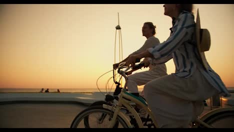 Side-view-of-a-happy-couple,-a-guy-in-light-clothes-and-a-girl-with-a-straw-hat-ride-bicycles-along-the-beach-near-the-sea-at-sunrise-in-summer.-A-guy-and-a-girl-are-driving-along-the-beach-at-dawn