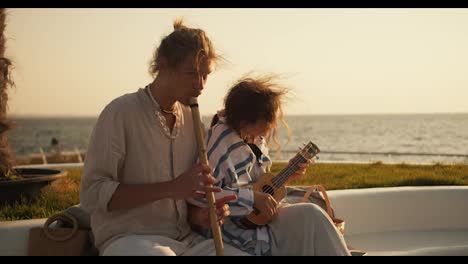 Fröhliches-Musikduo.-Ein-Typ-Spielt-Flöte,-Während-Das-Mädchen-Am-Strand-In-Der-Nähe-Des-Meeres-Ukulele-Spielt.-Ein-Typ-Spielt-Oboe-Am-Strand-In-Der-Nähe-Des-Meeres.-Ein-Blonder-Typ-In-Heller-Kleidung-Spielt-Ein-Blasinstrument-Aus-Bambus-Am-Strand-In-Der-Nähe-Des-Meeres.