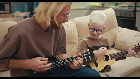 Un-Hombre-Rubio-Feliz-Con-Su-Pequeño-Hijo-Albino,-Con-Gafas-Azules,-Tocando-El-Ukelele-En-Un-Sofá-De-Color-Crema-En-Un-Apartamento-Moderno-Por-La-Mañana.-Un-Hombre-Rubio-Feliz-Enseñando-A-Su-Pequeño-Hijo-A-Tocar-La-Guitarra-Correctamente.