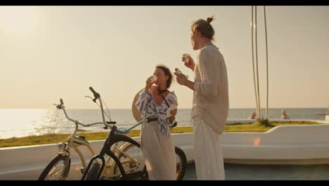 Happy-date-guy-together-with-his-girlfriend-drinking-coffee-and-eating-hot-dogs-near-their-bikes-on-a-modern-beach-near-the-sea-in-the-morning