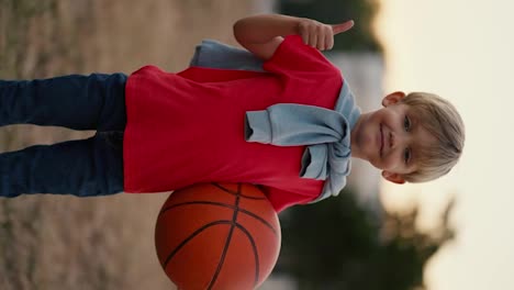 Vertical-video-portrait-of-a-happy-little-blond-boy-with-blue-eyes-showing-a-like-sign-and-sticking-his-thumb-up-while-standing-with-a-basketball-in-his-hands.-Happy-little-boy-has-fun-spending-his-leisure-time-actively-playing-sports-games