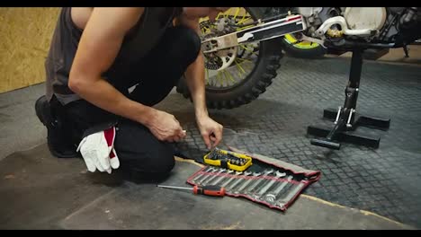 Primer-Plano:-Un-Hombre-Con-Una-Camiseta-Gris-Repara-E-Inspecciona-Su-Motocicleta,-Que-Levantó-Con-Un-Gato-En-Un-Taller-De-Garaje.-Un-Mecánico-Selecciona-La-Herramienta-Que-Necesita-De-Un-Conjunto-Completo-De-Herramientas.